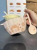 Hand holding a white ceramic spoon with cute bear and donut design next to a white ceramic bowl with matching bear and donut pattern on a dark surface.