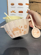 Hand holding a white ceramic spoon with cute bear and donut design next to a white ceramic bowl with matching bear and donut pattern on a dark surface.