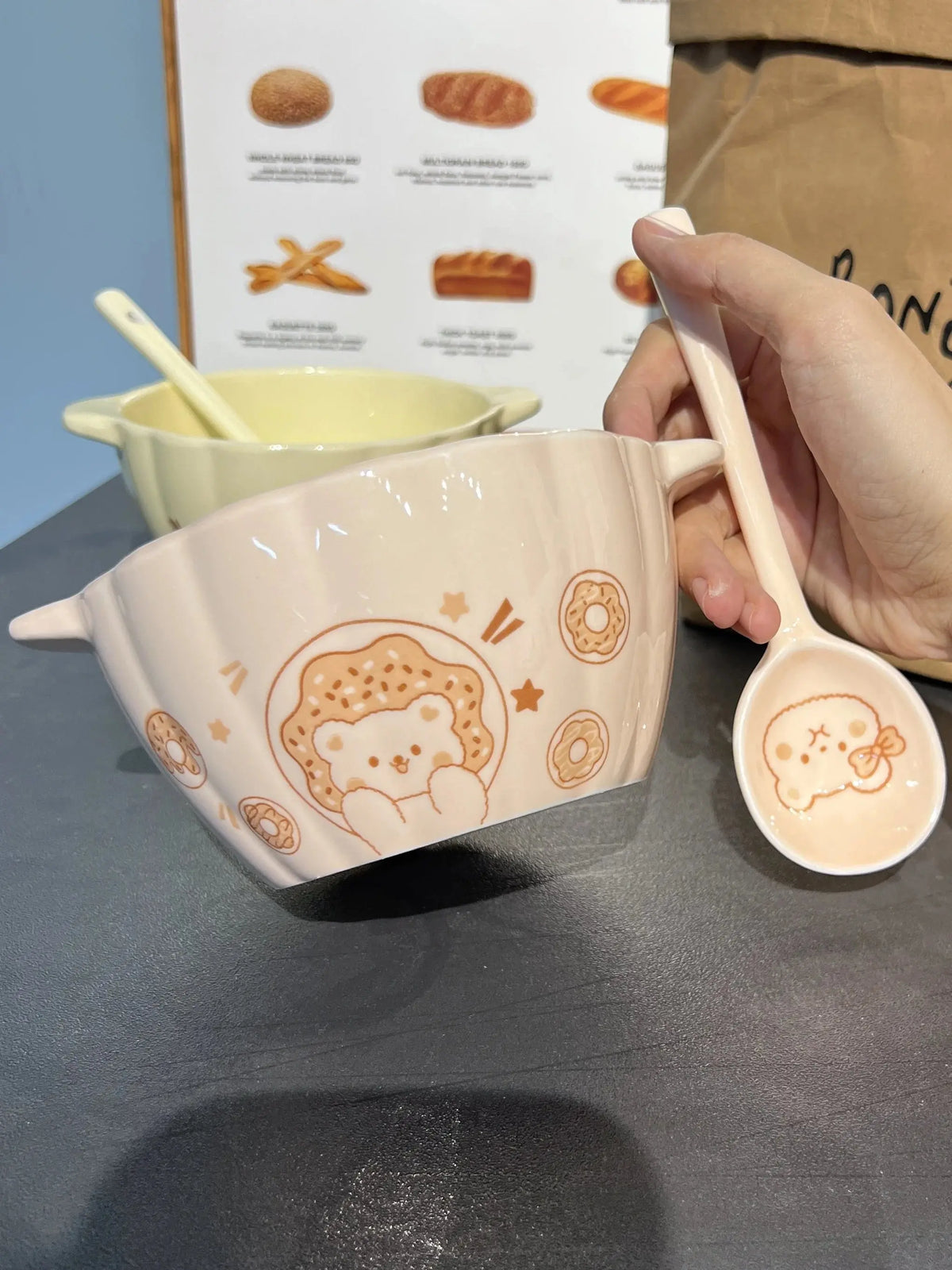 Hand holding a white ceramic spoon with cute bear and donut design next to a white ceramic bowl with matching bear and donut pattern on a dark surface.