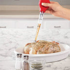 Hand holding red meat injector inserting seasoning into roast in a white dish